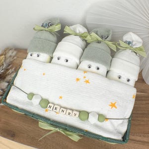 Könnte beinhalten: Ein grüner und weißer Baby-Geschenk-Korb mit einer weißen Decke, vier Sockenpuppen und einer Holzperlenkette mit dem Namen "HENRY" geschrieben.