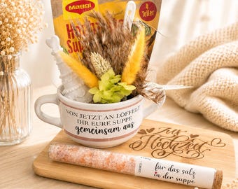 May include: A gift set featuring a white mug with text, dried flowers, and a packet of soup mix. A wooden cutting board with text and a tube of pink salt complete the arrangement. The mug reads "VON JETZT AN LÖFFELT IHR EURE SUPPE gemeinsam aus."