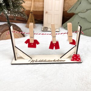 Puede incluir: Un tendedero de ropa de madera en miniatura con el texto "Wichtelwäscheleine" en la base. Prendas de ropa de fieltro rojo cuelgan de la línea con pinzas de madera.