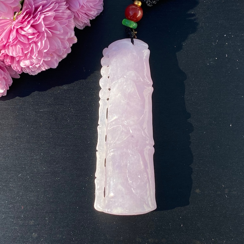 Puede incluir: Colgante de jade de color p&uacute;rpura claro con detalles de relieve tallados, suspendido de un cord&oacute;n negro con detalles rojos, verdes y dorados. El colgante es rectangular con bordes redondeados. Flores rosas en el fondo.