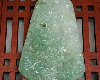 Op de afbeelding: Een doorschijnende, lichtgroene jade hanger met een gebeeldhouwd drakenontwerp. De hanger is rechthoekig met afgeronde randen en een getextureerd oppervlak. Een groen koord is aan de bovenkant bevestigd. Gepresenteerd op een houten oppervlak.