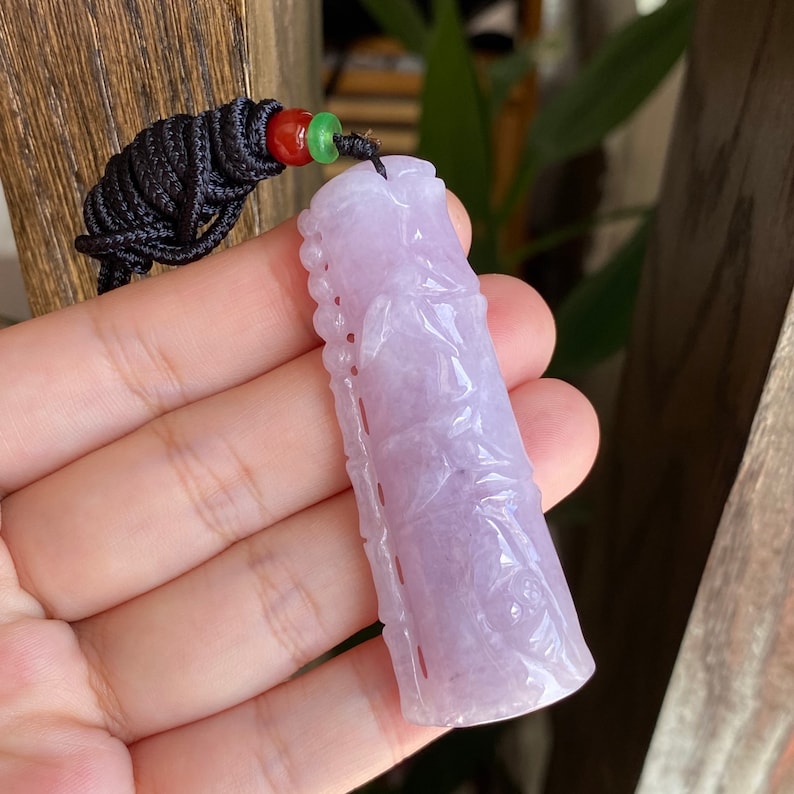 Puede incluir: Un colgante de jade lavanda con intrincadas tallas, suspendido de un cord&oacute;n negro con cuentas rojas y verdes. El colgante rectangular presenta patrones en relieve detallados. El fondo est&aacute; borroso.