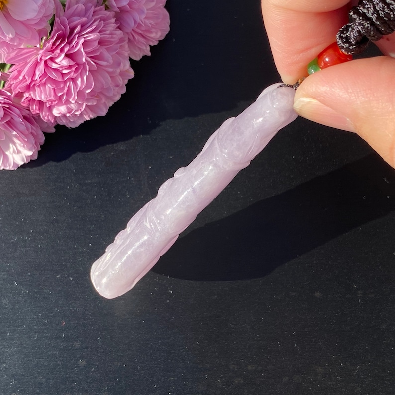 Puede incluir: Un colgante de jade tallado de color lavanda con detalles intrincados. El colgante est&aacute; sujeto por un cord&oacute;n negro con cuentas rojas y verdes. Flores rosas en el fondo. El colgante mide aproximadamente 10 cm de largo.