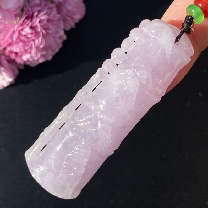Puede incluir: Un colgante de jade de color lavanda tallado con un dise&ntilde;o de bamb&uacute;. El colgante est&aacute; suspendido de un cord&oacute;n negro con cuentas rojas y verdes. El tallado detalla los tallos y las hojas de bamb&uacute;. El fondo es una superficie oscura con flores rosas.