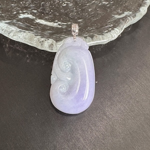 Könnte beinhalten: Ein lavendel-grüner Jade-Anhänger mit einer silbernen Öse. Der Anhänger hat ein wirbelndes, organisches Design. Der polierte Stein ist glatt und hat eine durchscheinende Qualität. Der Anhänger wird auf einer dunklen Oberfläche präsentiert.