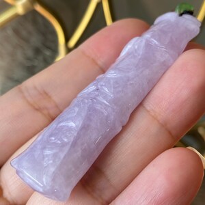 Puede incluir: Un colgante de jade lavanda con detalles florales tallados. El colgante es de color morado claro y tiene una superficie lisa y pulida. Una peque&ntilde;a cuenta verde est&aacute; unida en la parte superior. El colgante se sostiene en una mano.