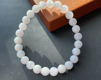 May include: A bracelet made of round, pale lavender beads. The beads have a slightly translucent appearance and are strung together on an elastic band. The bracelet is resting on a dark surface with a wooden accent.