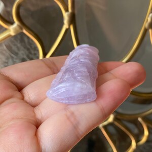 Puede incluir: Una figura de piedra tallada de color lavanda. La escultura tiene una superficie detallada y texturizada y parece ser una representaci&oacute;n estilizada de una figura. La piedra se sostiene en una mano, con un fondo borroso.