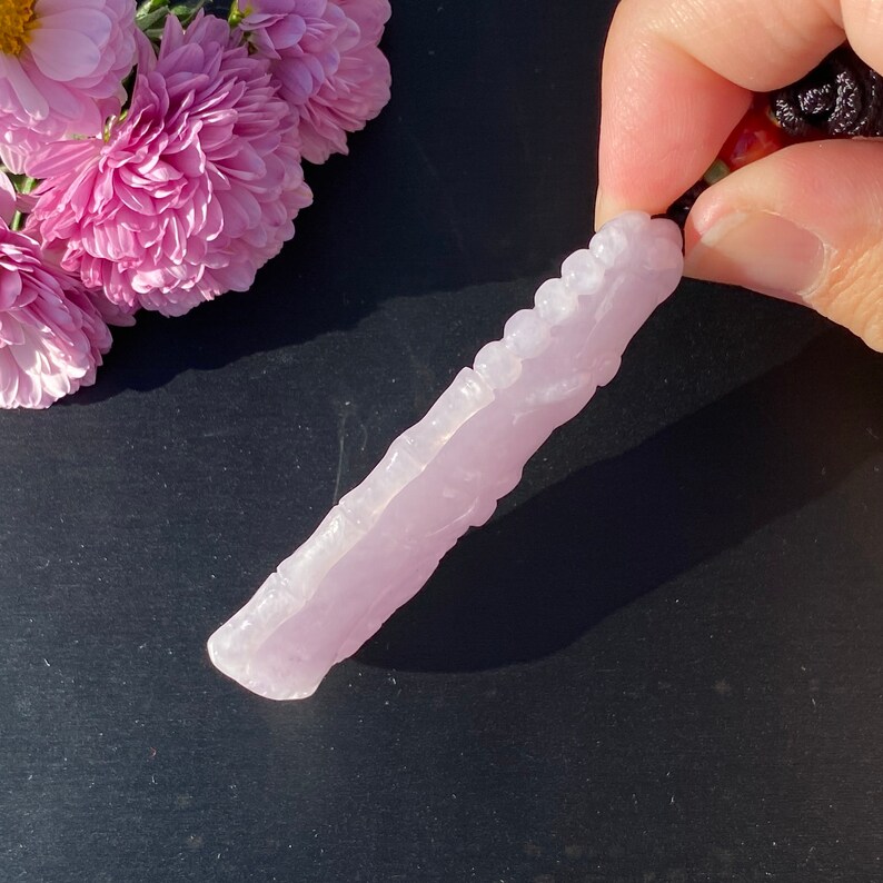 Puede incluir: Un colgante de cristal tallado de color rosa claro con una superficie texturizada. El colgante est&aacute; sujeto por un cord&oacute;n negro y se muestra sobre un fondo oscuro con flores rosas.