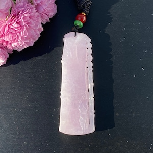 Puede incluir: Colgante de jade de color p&uacute;rpura claro con detalles de bamb&uacute; tallado. El colgante rectangular tiene una parte superior redondeada y un borde tallado. Cuelga de un cord&oacute;n negro con cuentas rojas y verdes. Flores rosas en el fondo.