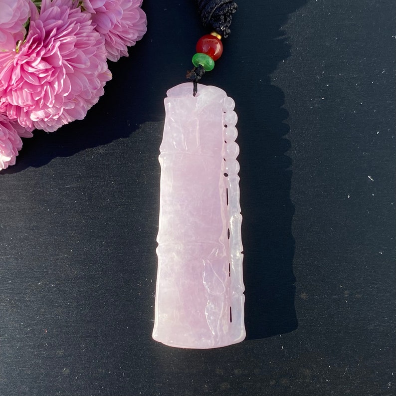 Puede incluir: Colgante de jade de color p&uacute;rpura claro con detalles de bamb&uacute; tallado. El colgante rectangular tiene una parte superior redondeada y un borde tallado. Cuelga de un cord&oacute;n negro con cuentas rojas y verdes. Flores rosas en el fondo.