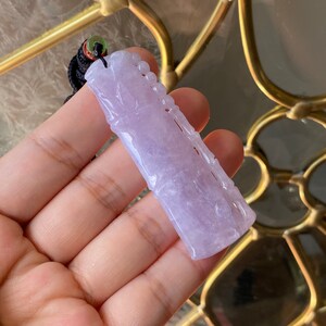 Puede incluir: Un colgante de jade color lavanda tallado con un dise&ntilde;o de bamb&uacute;. El colgante rectangular tiene detalles intrincados y cuelga de un cord&oacute;n negro con un acento verde.