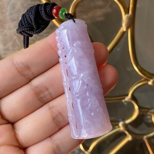 Puede incluir: Un colgante de jade lavanda con intrincadas tallas, suspendido de un cord&oacute;n negro con cuentas rojas y verdes. El colgante presenta un trabajo en relieve detallado y una superficie pulida, mostrando variaciones de color naturales.