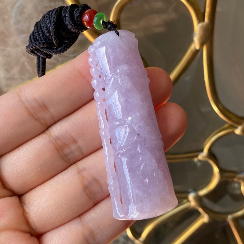 Puede incluir: Un colgante de jade lavanda con intrincadas tallas, suspendido de un cord&oacute;n negro con cuentas rojas y verdes. El colgante presenta un trabajo en relieve detallado y una superficie pulida, mostrando variaciones de color naturales.