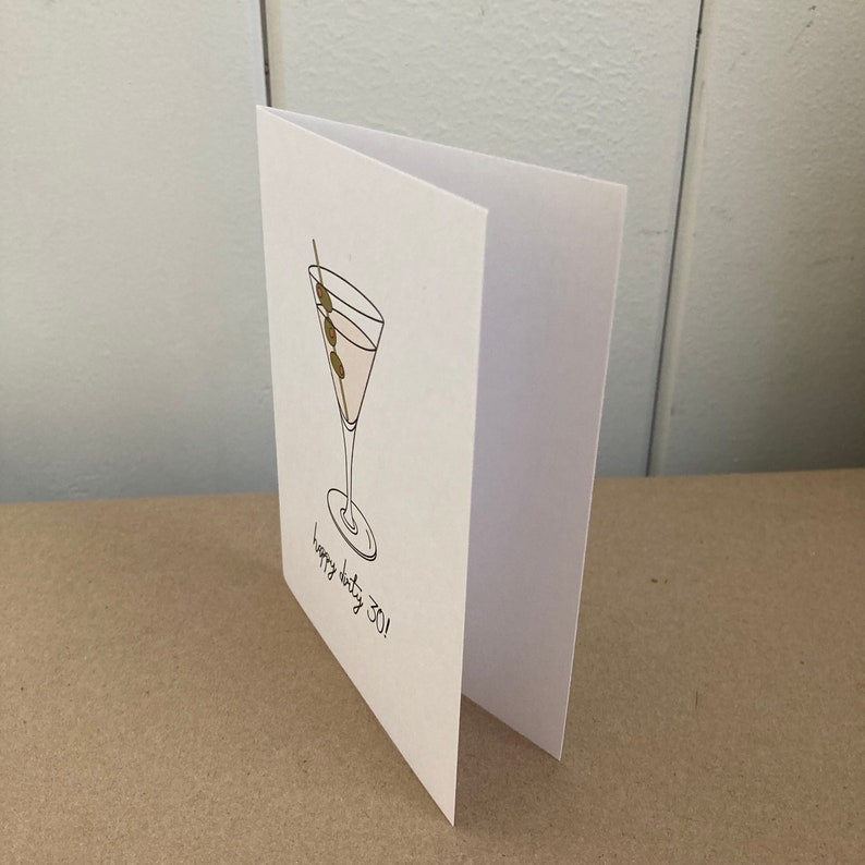 May include: A white card with a black line drawing of a martini glass with three olives on a toothpick. The text "Happy dirty 30!" is written below the glass.