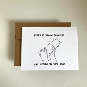 May include: A white card with black text and a line drawing of a cocktail shaker being shaken. The text reads "Here's to shaking things up and stirring up some fun!"
