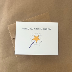 May include: A white birthday card with the text "WISHING YOU A MAGICAL BIRTHDAY!" and a yellow star wand design. The card rests on a brown envelope, against a brown backdrop.