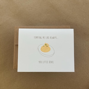 May include: A white greeting card with a cartoon illustration of a deviled egg. The text on the card reads "Tempting me like always... You little devil." The card is inside a brown envelope.