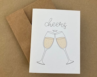 May include: A white card with the word "cheers" written in black cursive script. Two champagne glasses clink together, filled with a light beige liquid.