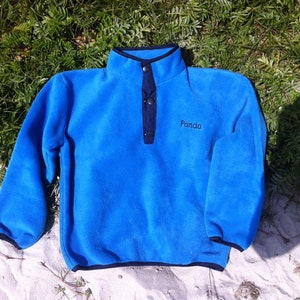 Peut inclure: Un pull en polaire bleu avec une fermeture à bouton pression noire et un col bleu marine. La polaire a le mot "Panda" brodé sur le devant.