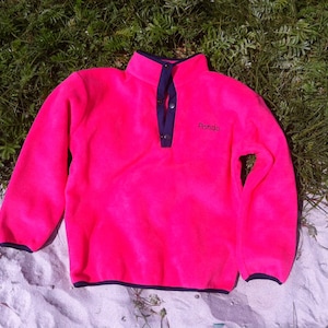 Könnte beinhalten: Ein leuchtend pinkfarbener Fleece-Pullover mit dunkelblauem Saum und einem einzigen Knopfverschluss. Der Fleece hat das Wort "Pando" auf der Vorderseite gestickt.