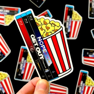 May include: A sticker featuring a popcorn bucket with red and white stripes, filled with popcorn and three Blu-ray movie cases. The movie titles are "Us", "Get Out", and "Nope".