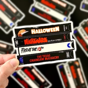 May include: A stack of four VHS tape stickers featuring classic horror movie titles: "Halloween," "Nightmare on Elm Street," "Friday the 13th," and "The Texas Chainsaw Massacre." The stickers are black with white and red text.