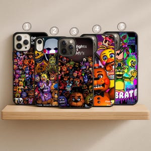 May include: Five phone cases with black borders are displayed on a wooden shelf. Each case features a colorful graphic design with characters and text from the video game "Five Nights at Freddy's". The text "lights t Idy's" is visible on one case.
