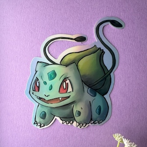 Op de afbeelding: Een holografische sticker met een cartoonillustratie van een groene Bulbasaur Pokémon met een rood oog en een groen blad op zijn rug.