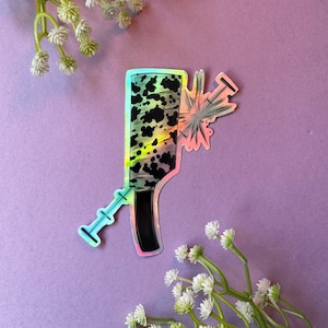 Puede incluir: Una pegatina hologr&aacute;fica de un cuchillo de carnicero con estampado en blanco y negro con una jeringa que sobresale de la hoja.