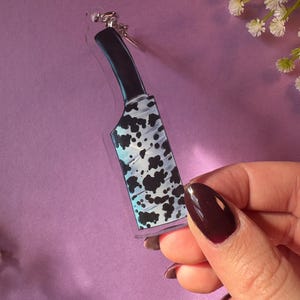 May include: A clear acrylic keychain with a black and white cow print design. The keychain is shaped like a knife.