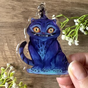 May include: A blue cat-shaped keyring with a cartoon design. The cat has yellow eyes, sharp teeth, and a tail. The keyring is made of clear plastic and has a silver-coloured metal clasp.