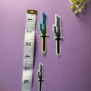 May include: Three stickers of pixelated swords with a rainbow gradient. The swords are on a purple background with a ruler for scale.