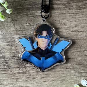 Peut inclure: Un porte-clés en acrylique transparent représentant un personnage de super-héros avec des cheveux noirs, un masque bleu et un costume bleu et noir. Le porte-clés a un fermoir noir et un motif d'emblème bleu. Le fond est une surface en bois.