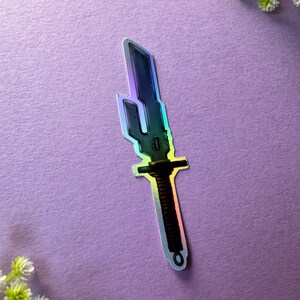 May include: A holographic sticker of a silver and brown sword with a detailed design. The sword has a long, thin blade and a rounded hilt.