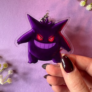 May include: A clear acrylic keychain featuring a purple and black cartoon ghost with red eyes. The ghost has a menacing grin and is in a standing pose.