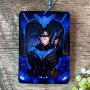 Peut inclure: Un porte-clés rectangulaire sur le thème de Nightwing, avec une illustration stylisée du personnage en costume bleu et noir. Le design comprend une toile de fond de paysage urbain, des accents floraux bleus et une chaîne de perles bleues.
