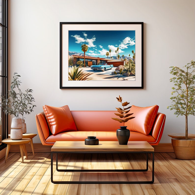 May include: A framed print of a mid-century modern home with a blue car parked in the driveway. The home is surrounded by palm trees and desert landscaping. The print is hanging on a white wall in a living room setting with an orange couch and a coffee table.