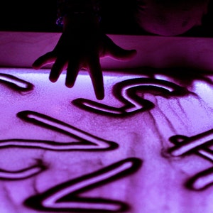 May include: A hand draws numbers in a light purple sand tray. The numbers are 1, 2, 3, 4, 5, 6, 7, and 8.