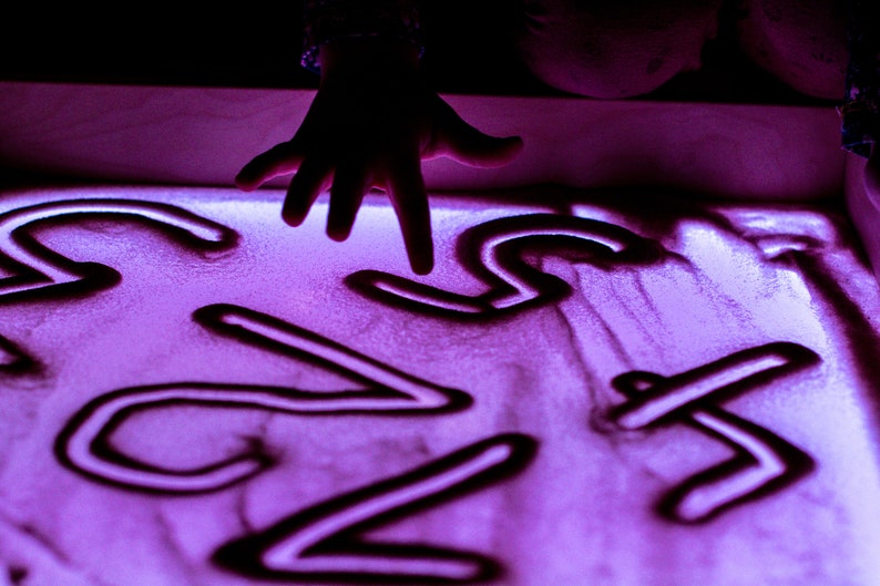 May include: A hand draws numbers in a light purple sand tray. The numbers are 1, 2, 3, 4, 5, 6, 7, and 8.