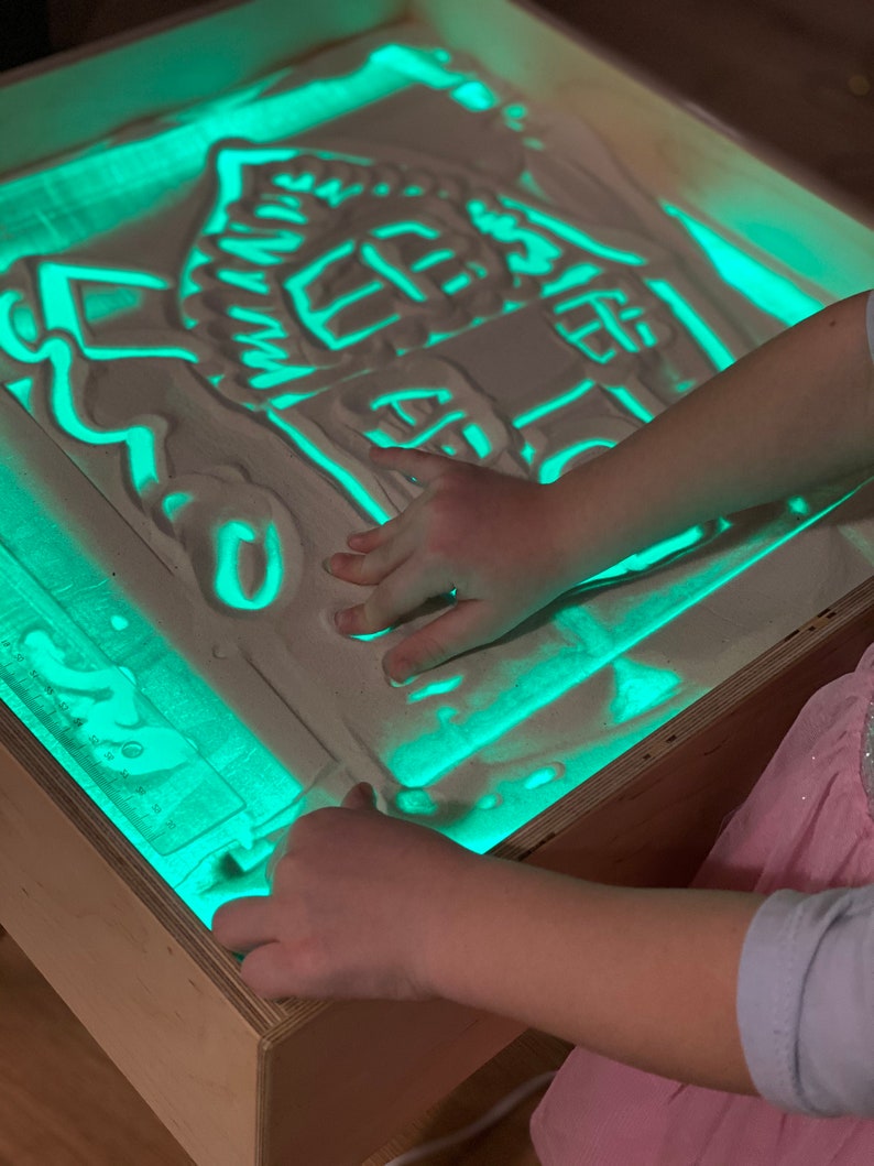 May include: A child's hand is drawing a house shape in a light-up sand table. The sand table is made of wood and has a green light shining through the sand.