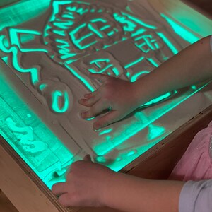 May include: A child's hand is drawing a house shape in a light-up sand table. The sand table is made of wood and has a green light shining through the sand.