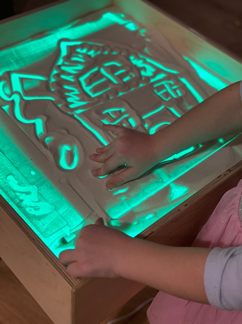 May include: A child's hand is drawing a house shape in a light-up sand table. The sand table is made of wood and has a green light shining through the sand.