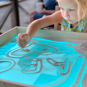 May include: A child uses their finger to draw shapes in a light blue sand tray with white sand. The tray is illuminated from below, creating a glowing effect.