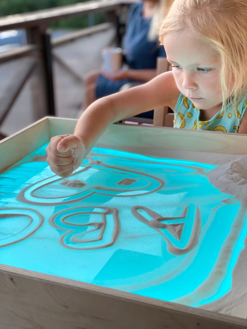 May include: A child uses their finger to draw shapes in a light blue sand tray with white sand. The tray is illuminated from below, creating a glowing effect.