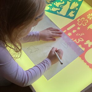 May include: A child uses a light table to trace animal stencils with a pencil. The light table has a yellow surface and is surrounded by a wooden frame. The stencils are colorful and feature various animals, including a cat, a dog, a bird, and a fish.