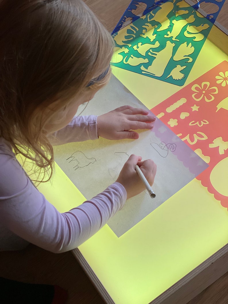 May include: A child uses a light table to trace animal stencils with a pencil. The light table has a yellow surface and is surrounded by a wooden frame. The stencils are colorful and feature various animals, including a cat, a dog, a bird, and a fish.