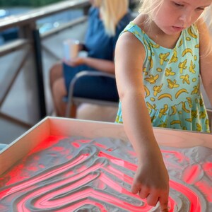 May include: A young girl draws a red heart shape in a light sand tray with her finger. The tray is lit from below, making the lines glow red.