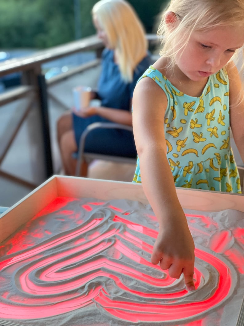 May include: A young girl draws a red heart shape in a light sand tray with her finger. The tray is lit from below, making the lines glow red.