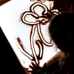 May include: A child's hand draws a flower design in the sand on a light table.